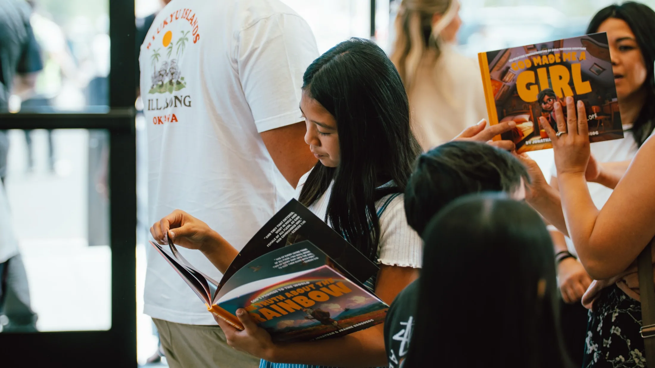 Readers at a church book table, one girl reading an open copy of The Truth About The Rainbow while another holds up God Made Me A Girl